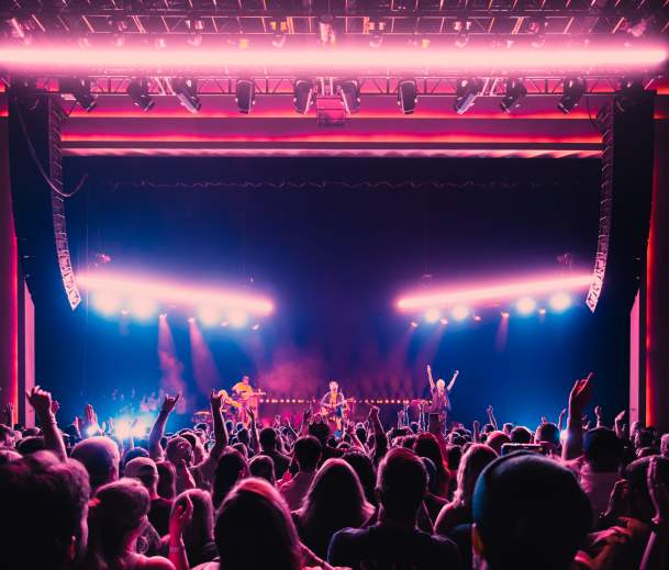 Crowd at a concert at Channel 24, with a band performing on stage under bright pink and white lights as audience members raise their hands and phones.