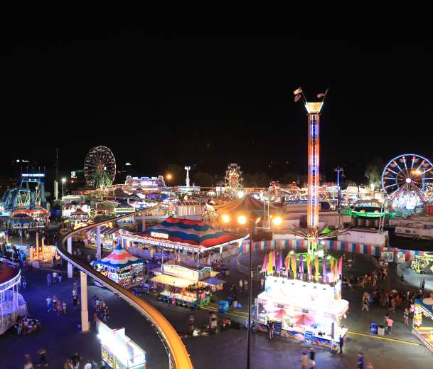 A nighttime photo of the state fair skyline, with neon lit rides including ferris wheels and drop rides