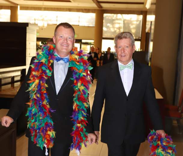 Two smiling gala attendees in tuxedos with rainbow feather boas