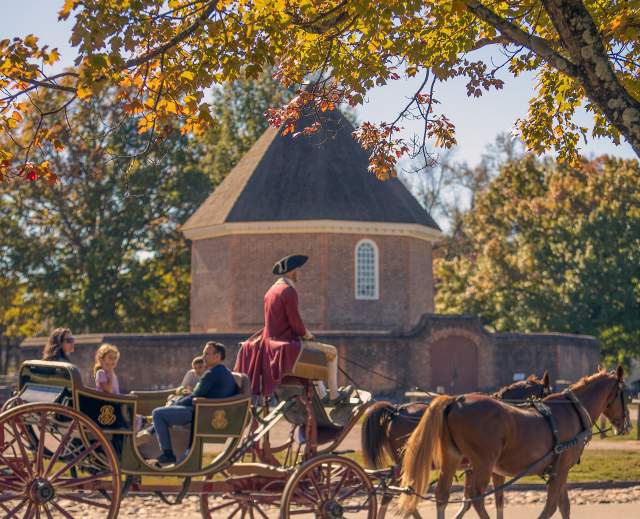 A horse-drawn carriage passes under colorful autumn leaves, with a historic round building in the background, evoking a charming, nostalgic atmosphere.