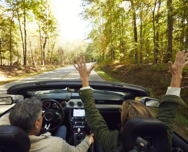 Couple driving convertible down nature filled road with roof down, and passenger has hands up