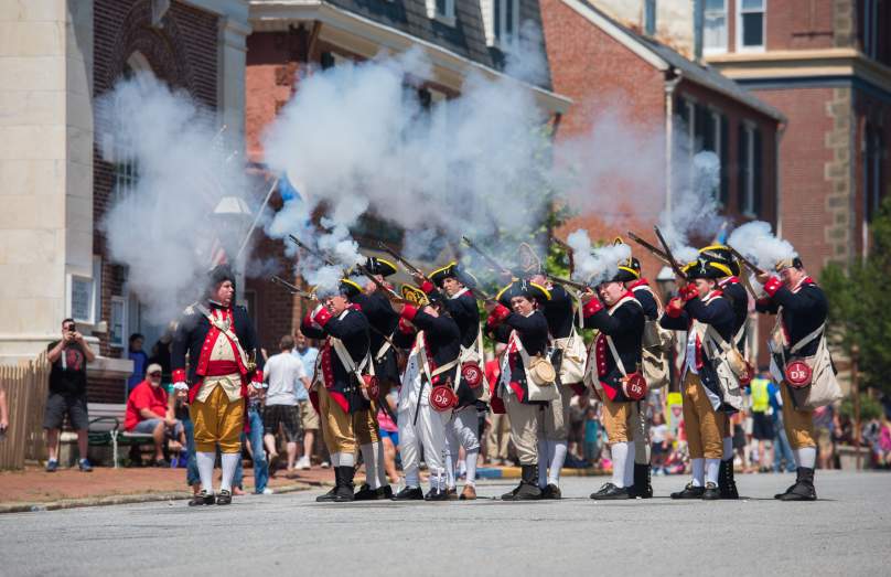 A group of historical reenactors in colonial military uniforms fires muskets, creating clouds of smoke during a lively outdoor event.