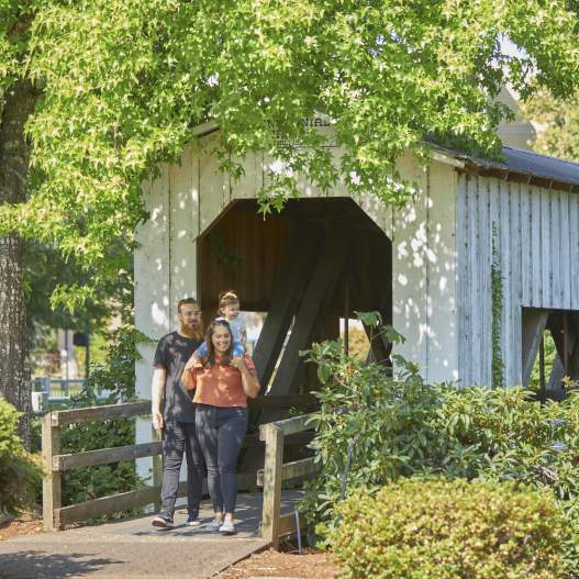 A young family of three exit the white, wooden Centennial Covered Bridge with trees overhead.