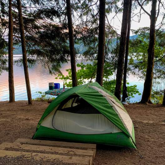 A green and white tent sits beside a lake with tall trees and sunrise colors reflecting off the water.