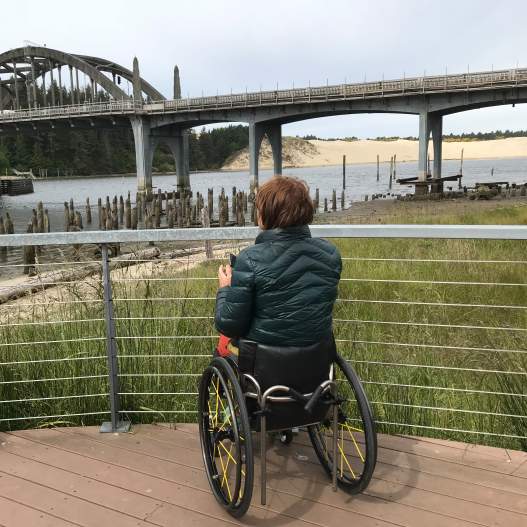 A person in a dark puffy coat with a wheelchair is on the flat boarded viewing point looking out toward the historic Siuslaw River Bridge. The viewing platform has a thin rail and wire fence allowing optimal viewing with the top rail below the person's sightline. Green rushes and grass fill in the space between the deck and riverside. Sand dunes are in the background.
