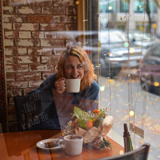 looking through a window at a woman smiling with a mug of coffee. there are flowers on her table and fairy lights in the background.