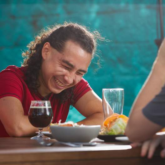 A man sits at a table with food and a dark beer. He is laughing with friends at the edge of the frame.