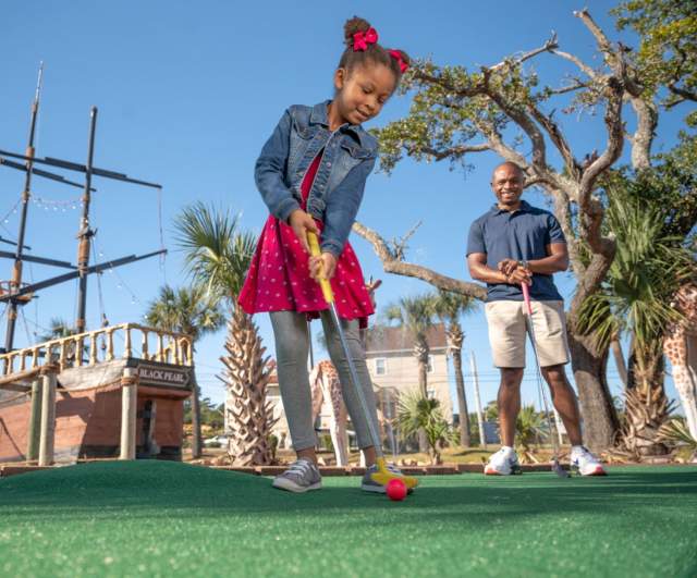 a little girl putting at a mini golf course while her dad looks on.