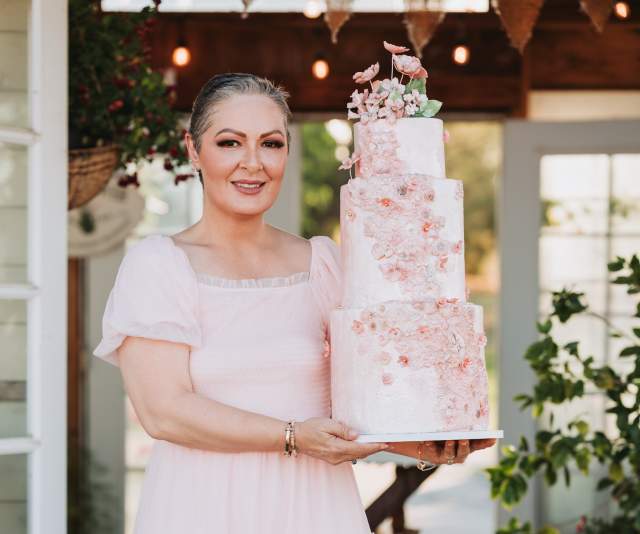 A chef in a soft pink dress proudly displays a three-tier pink floral cake