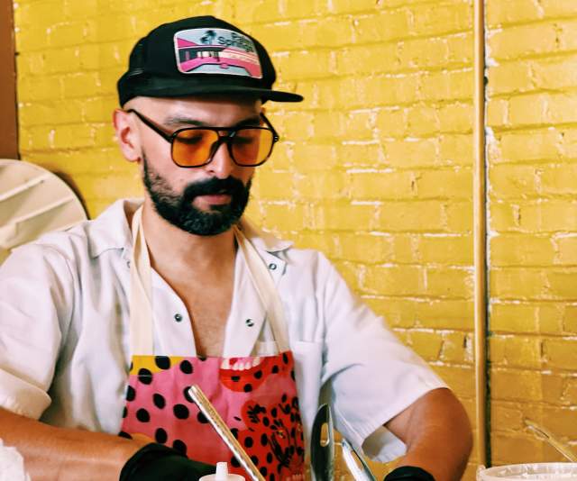 A chef in a white overcoat, bright pink floral apron, sunglasses, and ballcap in front of a yellow brick wall