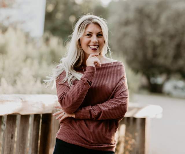 A woman in a dusty maroon sweater smiles in front of a rustic fence and tree-lined landscape
