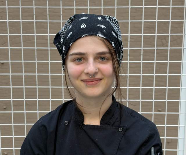 A chef in a black overcoat and bandana smiles with arms crossed in front of a brown tiled wall