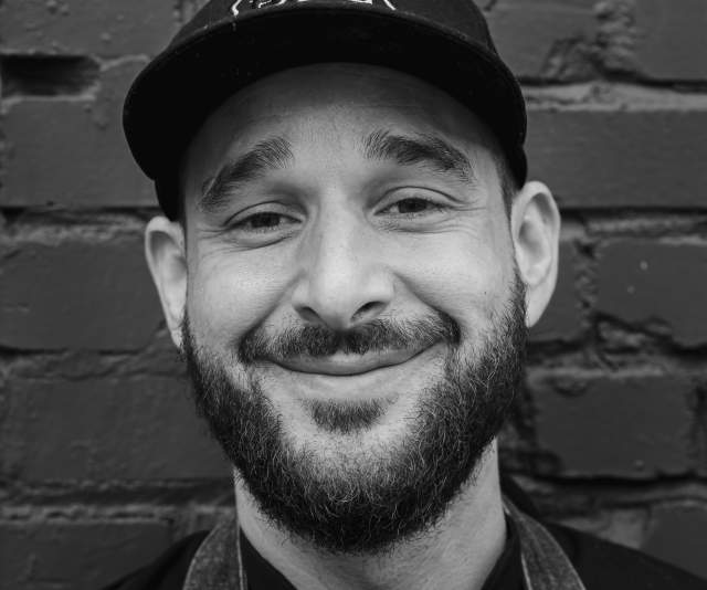 A black and white photo of a chef in a dark shirt and dark ballcap smiling in front of a brick wall