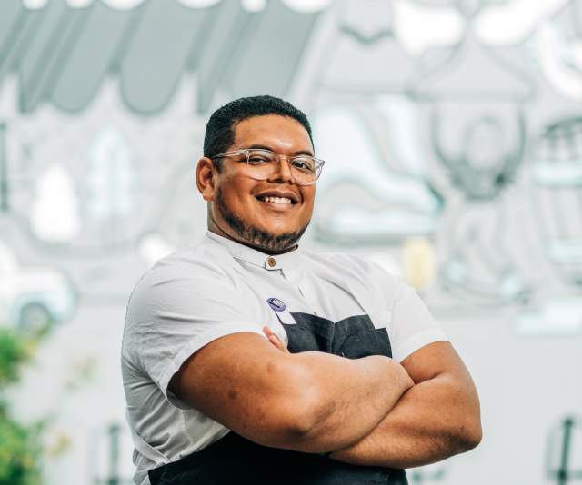 A chef in a light gray t-shirt and black apron smiles and stands with arms crossed in front of a dusty blue mural of "Urban Roots"