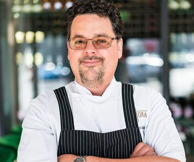 A chef in a white shirt and pinstriped black apron smiles with arms crossed in front of a bar in a sunlit restaurant