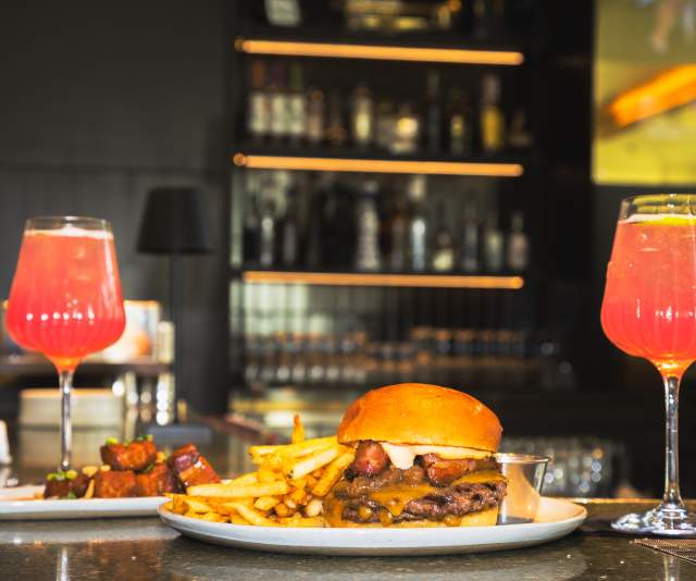 Burger and fries from The 7th Street Standard served alongside two bright pink spritz cocktails at a dimly lit bar.