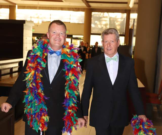Two smiling gala attendees in tuxedos with rainbow feather boas
