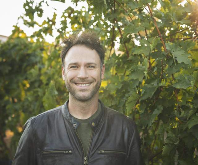A winemaker in a green shirt and black leather jacket smiles in front of a row of grape vines