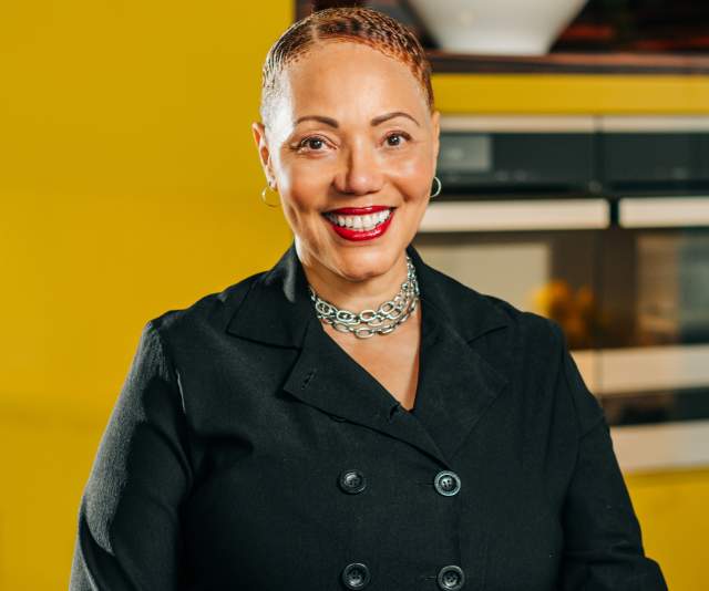 A chef in a black overcoat smiles in front of a cheery yellow wall