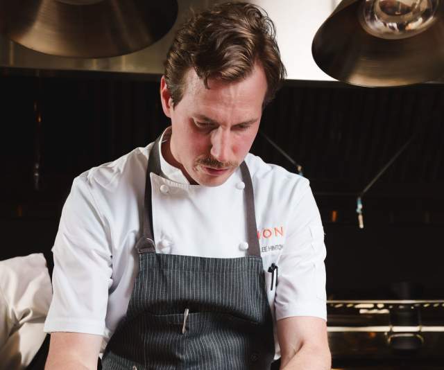 A chef in a white overcoat and black pinstripe apron artfully plates food in a restaurant kitchen