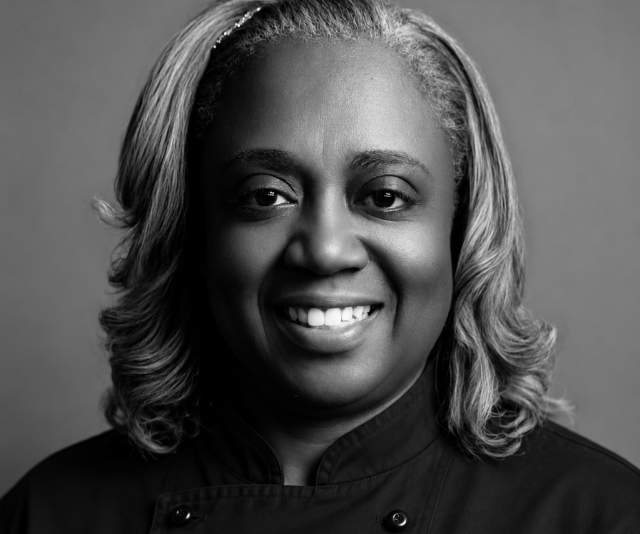 A black and white portrait of a smiling chef in a black overcoat
