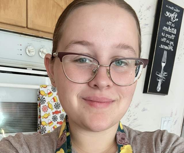 A smiling chef in a beige shirt, floral apron, and cat-eye glasses