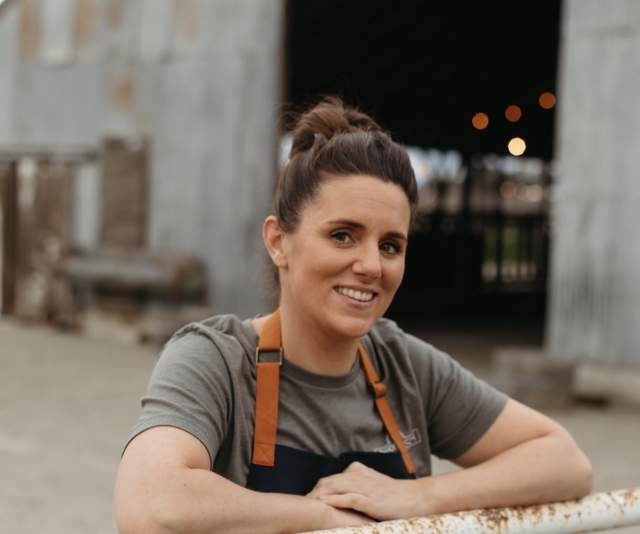 A chef in a black apron with orange straps smiles and leans against a fence in front of a barn