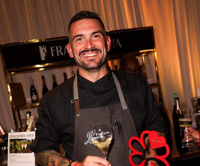 A chef in a black shirt and gray apron smiles and presents a Michelin award while toasting with a glass of wine