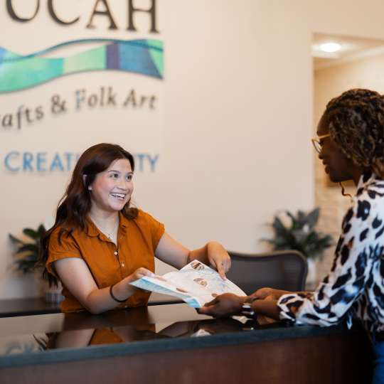 A woman stands behind the counter at the Paducah Convention & Visitors Bureau and helps another woman who is visiting Paducah. They are holding a map and pointing at a location.