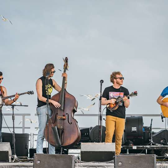 A band of four people—one with a banjo, cello, ukelele, and guitar—stands on stage on the beach.