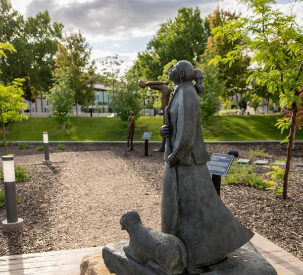 A dark metal sculpture of a woman, with a lamb at her feet, stands with her head tilted hopefully toward the sky on a slab of sandy stone.