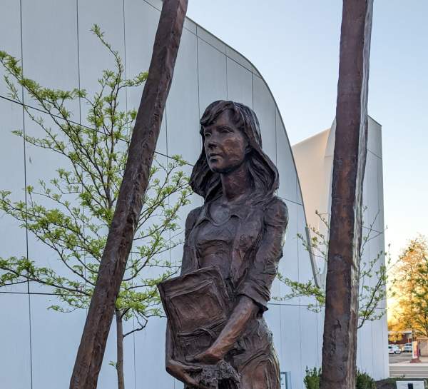 A bronze-cast statue of a woman in a dress, holding a book, stands in a geometric frame in front of a sweeping white building.