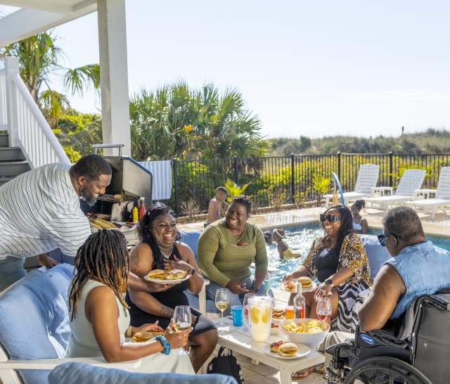 Multi-generational family enjoying a joyful reunion at a vacation rental in North Myrtle Beach, South Carolina, gathered around a poolside table with food and drinks, celebrating quality time together in a warm coastal setting.