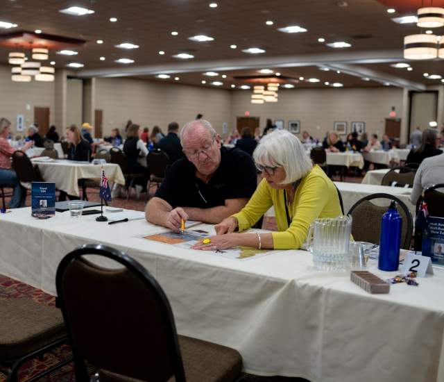 A man and a woman, sitting side by side at a table, look over a promotional item on the table in front of them in a conference room filled with other people at tables also in meetings.