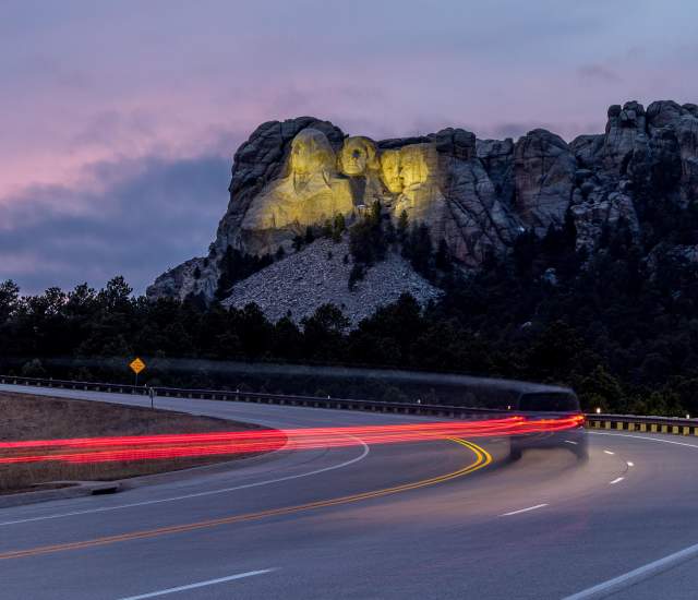 Mount Rushmore, lit up by yellow floodlights at dusk, sits in the background as a vehicle traveling down the highway is trailed by a long line of red taillights, created by a long-exposure photo.