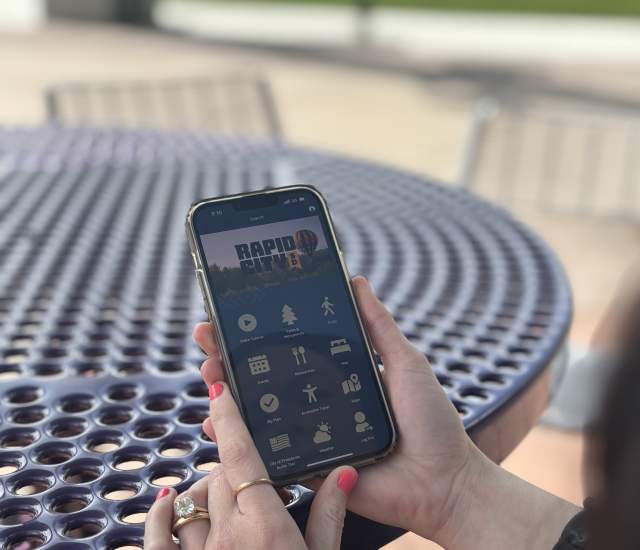 A woman holding her cell phone looks at the Visit Rapid City app while sitting at a picnic table