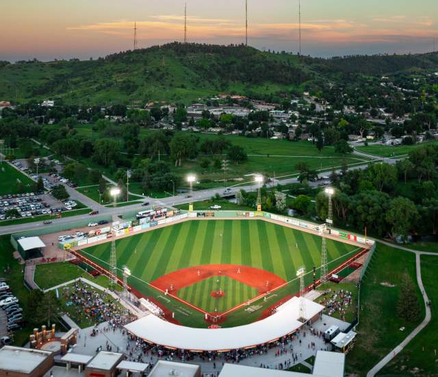 A high-angle view of Fitzgerald Stadium from behind home plate at dusk. The stadium lights illuminate the playing surface as the dusk sky darkens. The hills beyond center field are a vibrant green in peak summer.
