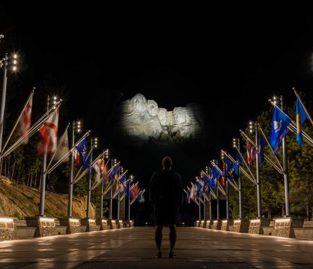 person standing at the avenue of flags looking up at an illuminated mount rushmore in the black hills of south dakota