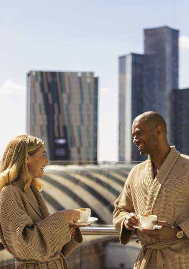 Couple on hotel balcony in dressign gowns drinking coffee with views over Manchester