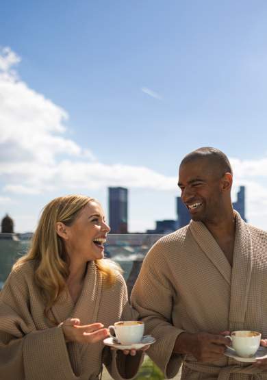 Couple in dressing gowns with cups of coffee stood on hotel balcony overlooking Manchester