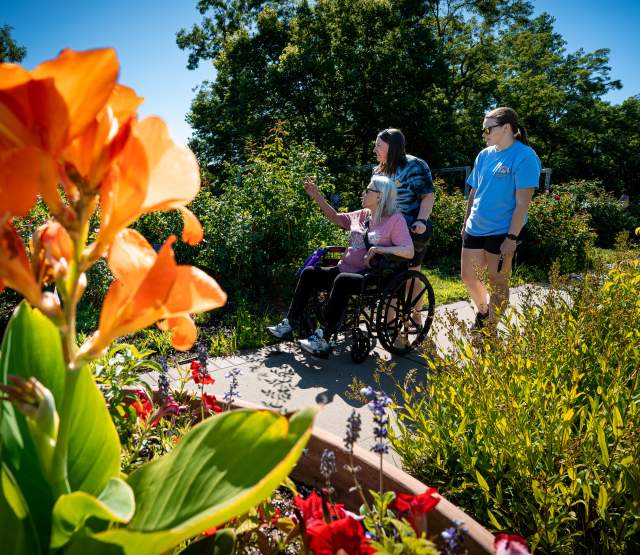 Three people, one in a wheelchair, enjoy a sunny Lauritzen Gardens walk with vibrant orange flowers in the foreground.