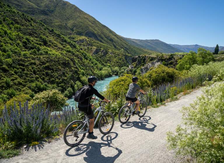 Two people biking along a blue river with wild flowers