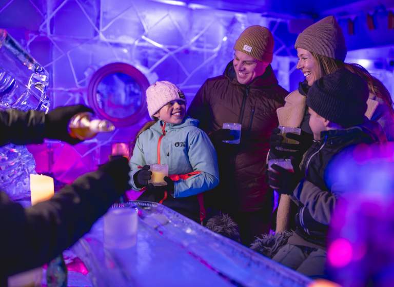 A family of four enjoying drinks in an ice bar, surrounded by illuminated ice sculptures and glowing purple and blue lighting. They are dressed warmly in coats, hats, and gloves, smiling and sharing a joyful moment.