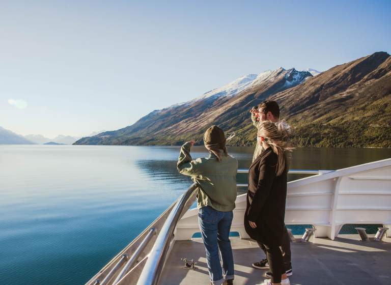 Three people standing on the deck of a boat, gazing out at the calm blue waters of a lake surrounded by mountains. The landscape features snow-capped peaks and lush greenery under a clear blue sky, creating a serene and picturesque setting