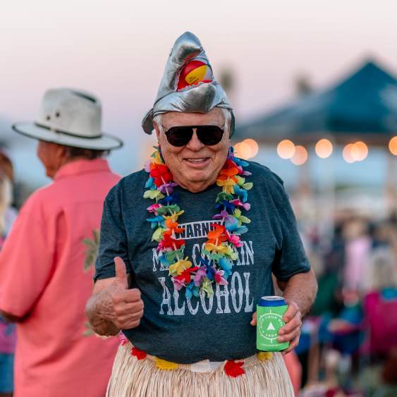 A man gives a thumbs up to the camera. He's wearing a metallic shark hat, a hula skirt, and several leis.
