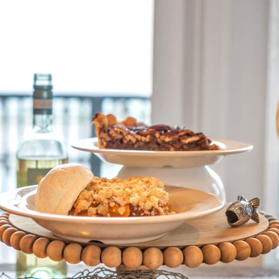A plate of cobbler with ice cream and a plate of pecan pie sit stacked on an elevated dessert tray. To the left are open bottles and glasses of wine.