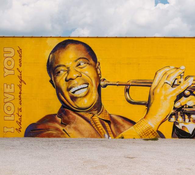 Photorealistic, bright yellow mural of Louis Armstrong smiling and holding his trumpet up to play. The words "I love you" and "What a wonderful world" are painted on the left hand side of the mural.