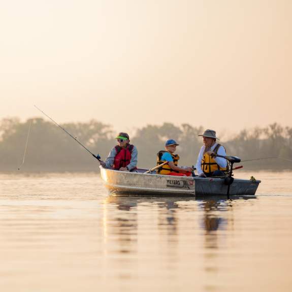 A canoe with three people paddles on calm water during sunset, surrounded by trees and a serene landscape. The scene evokes outdoor adventure.