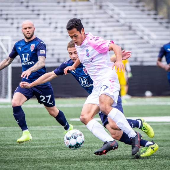 A Madison Forward soccer play dribbles the ball as two defenders from the other team approach him from the side.