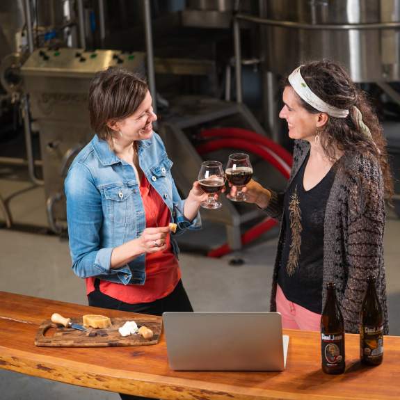 Two women standing in front of a laptop toast glasses of beer